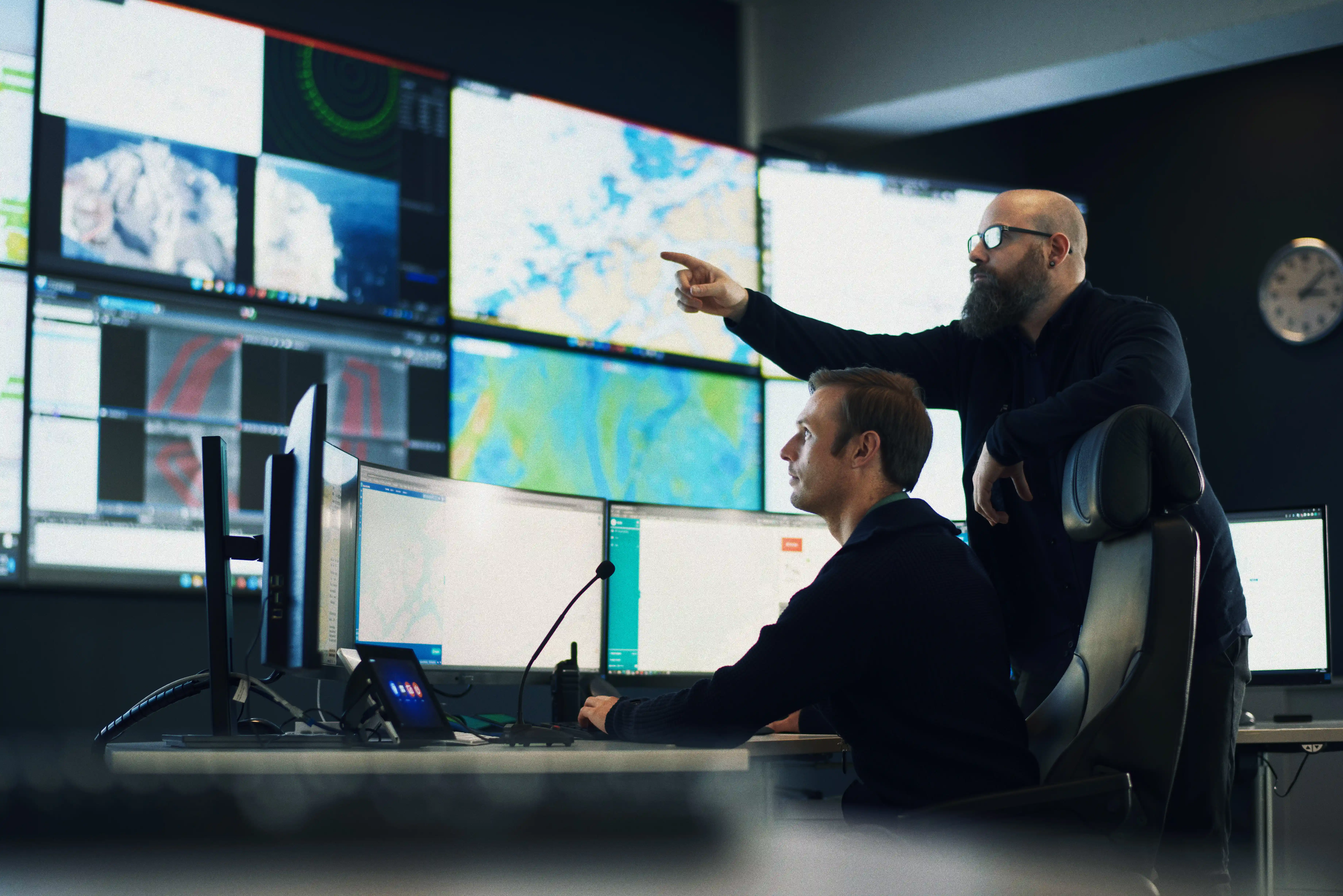Bildet viser to menn i Norseyes havovervåknings- og beredskapssentral. Bakveggen er dekt av store skjermer som viser områdene som overvåkes. En av de ansatte sitter ved en pult med monitorer foran seg, mens den andre mannen står bak og peker på de større skjermene.