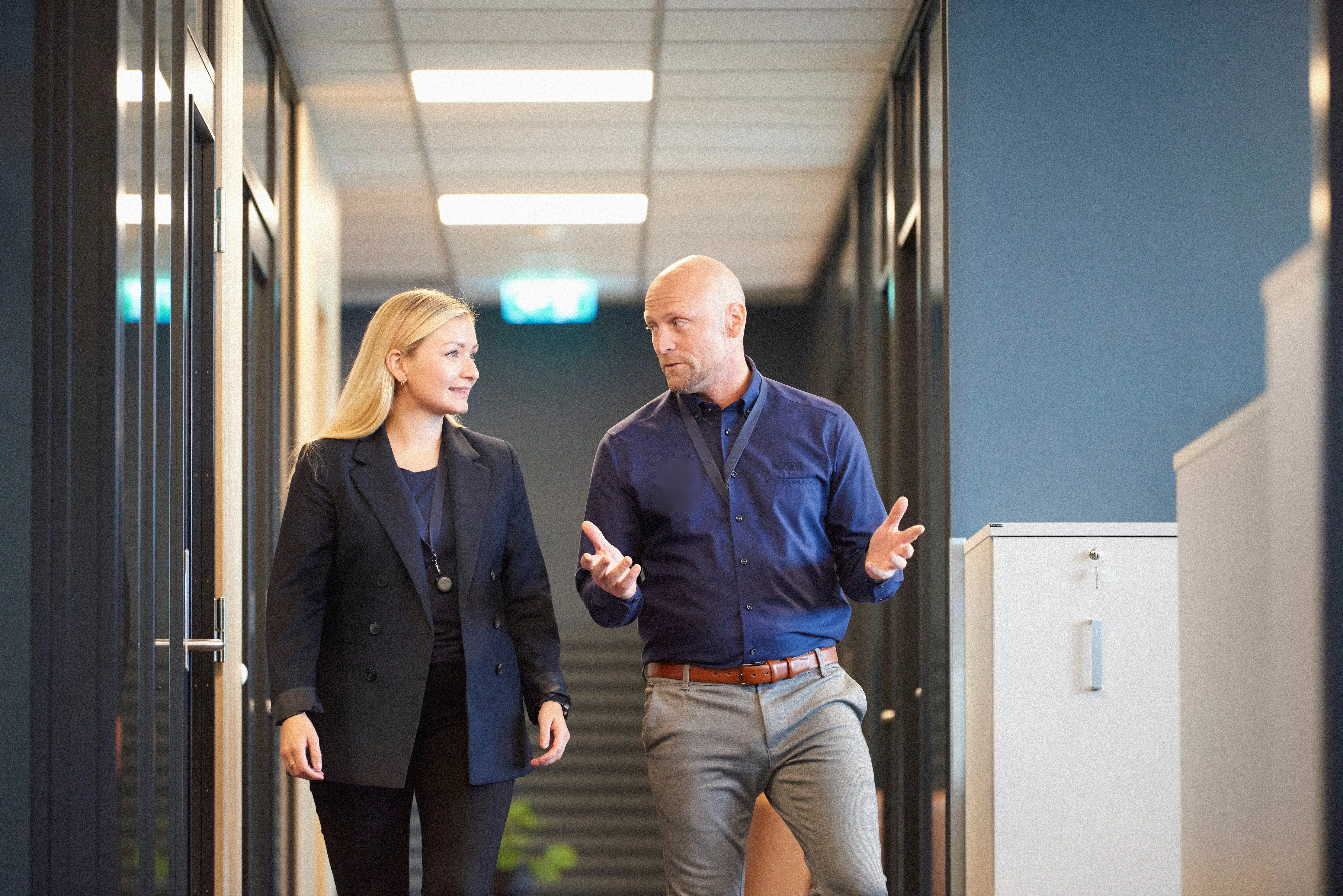 Bildet viser en kvinnelig og mannlig ansatt i dialog. De går nedover gangen på Norseye-kontoret.