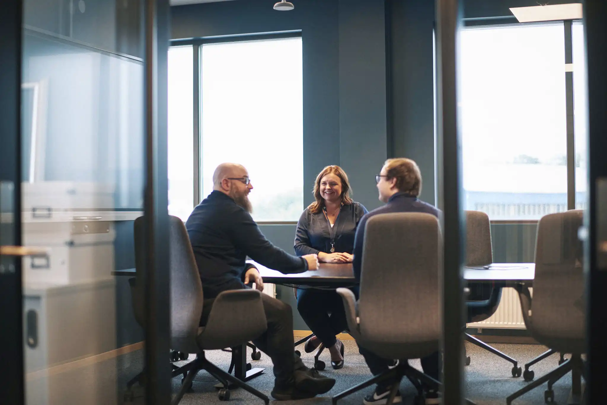 The image shows three employees in conversation at a table in Norseye’s meeting room. The photo was taken during the daytime, and there are two men and one woman. The employees are smiling at each other.