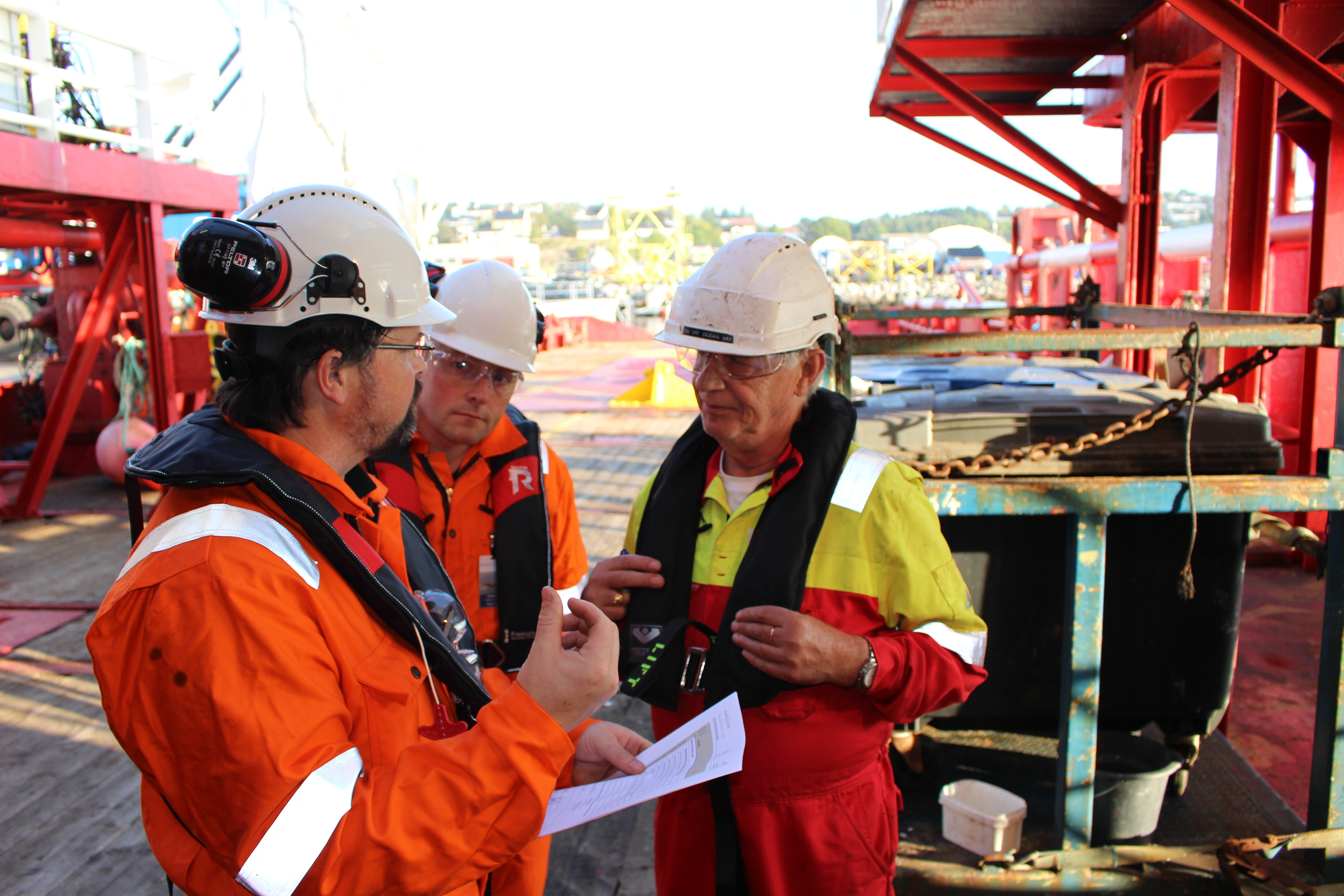 Bildet viser tre menn i arbeidstøy, vernebriller og hjelmer. En av dem holder papirer i hånden. De tre mennene er i dialog på en arbeidsplass. Bildet er tatt på dagtid.